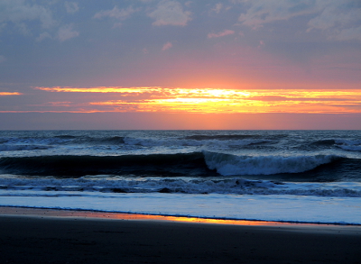 [A large wave curls toward the beach under the last of the violet orange gold setting sun.]